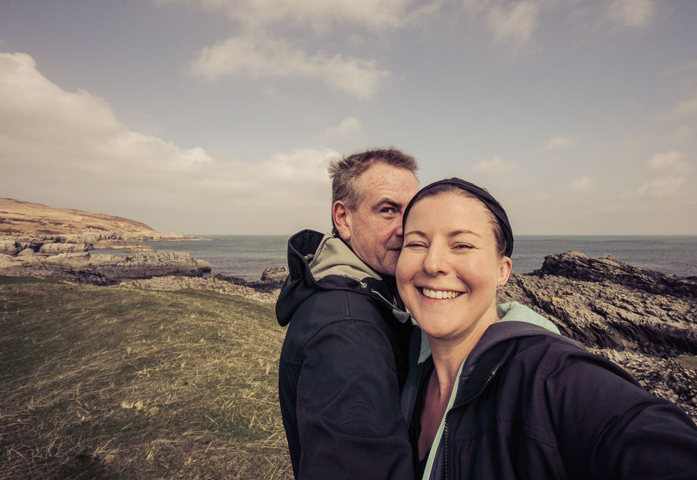 Photograph of Roddy MacEachern and Amanda Reid with the rocky shoreline of Sanaigmore Bay, Islay in the background. Roddy is trying to hide from the camera while Amanda is laughing at him.